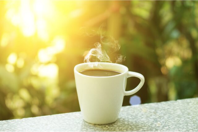 朝日を浴びながら湯気が立つコーヒーのカップ。日常に寄り添う香りのイメージ