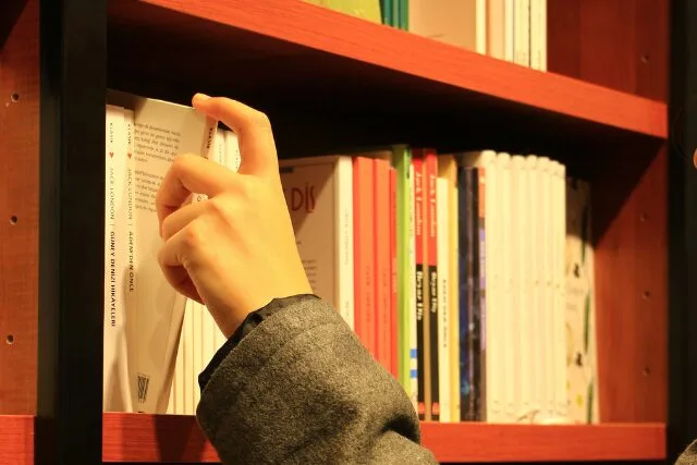 本棚の前で本を手に取る人の手元。やわらかな照明に照らされた書店の一角。