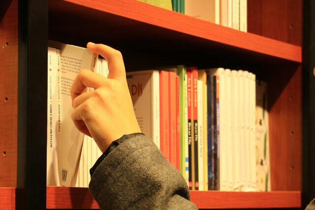 本棚の前で本を手に取る人の手元。やわらかな照明に照らされた書店の一角。