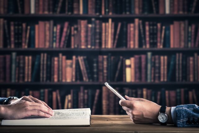 書店のテーブルで、本を開く手とスマートフォンを持つ手。デジタルとアナログの対比