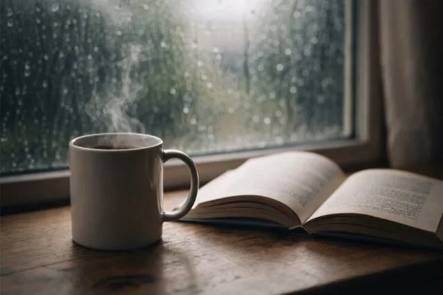 雨の窓辺にあるマグカップと本の写真